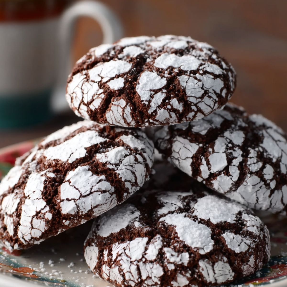 A plate with several chocolate crinkle cookies dusted with powdered sugar, with a cup of coffee in the background.