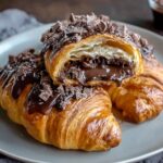 Close-up of a flaky chocolate croissants cut open to reveal rich melted chocolate filling, topped with dark chocolate drizzle and shavings on a gray plate.