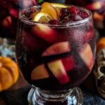 A close-up of chilled Halloween sangria with fruit chunks and orange slices, served in a frosty goblet, decorated with mini pumpkins and a skull.