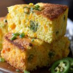 Two thick slices of jalapeño cheddar cornbread stacked on a wooden plate, showing melted cheese and green jalapeño bits, garnished with sliced jalapeños.