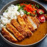 A bowl of Japanese katsu curry with crispy breaded chicken cutlets sliced neatly, served with white rice, rich brown curry sauce, and halved cherry tomatoes garnished with chopped herbs.