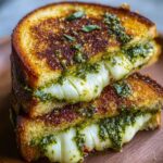 A close-up of a pesto grilled cheese sandwich cut in half, showing melted mozzarella and green pesto filling inside crispy, buttery bread.
