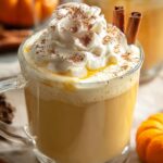 A clear glass mug filled with Pumpkin White Hot Chocolate chocolate, topped with whipped cream, cinnamon powder, and cinnamon sticks, surrounded by mini pumpkins and pinecones.