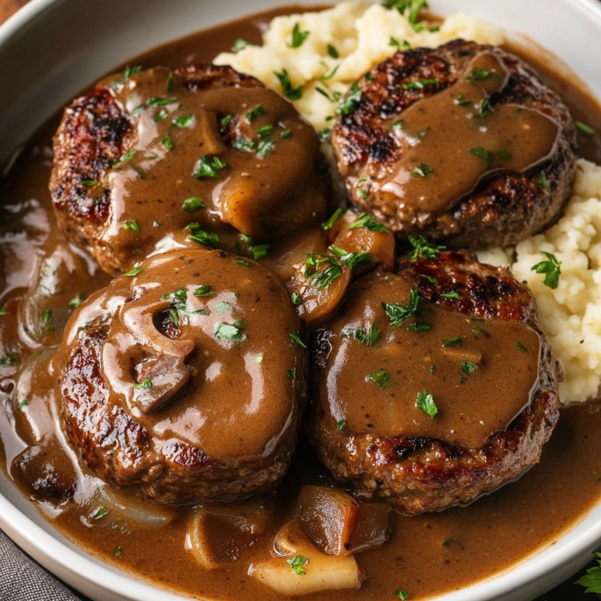 A delicious plate of Salisbury steak is served with mashed potatoes and a savory brown gravy. The steaks are topped with onions and the rich sauce, making for a comforting, hearty meal.
