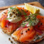 Two slices of toast topped with cream cheese, Smoked Salmon Breakfast, capers, red onions, fresh dill, and lemon wedge on a white plate.