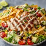 A vibrant Southwest-style Spicy Southwest Salad topped with grilled chicken slices, drizzled with creamy dressing. The salad includes black beans, corn, cherry tomatoes, avocado chunks, romaine lettuce, and crispy tortilla strips, all arranged on a ceramic plate.