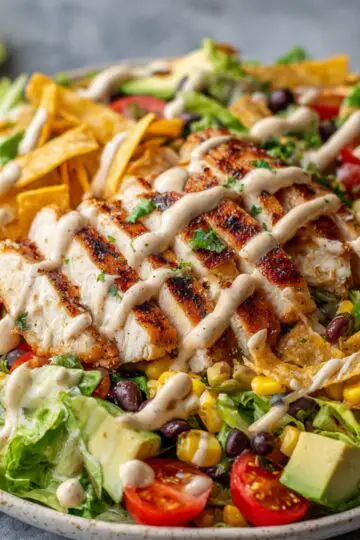A vibrant Southwest-style Spicy Southwest Salad topped with grilled chicken slices, drizzled with creamy dressing. The salad includes black beans, corn, cherry tomatoes, avocado chunks, romaine lettuce, and crispy tortilla strips, all arranged on a ceramic plate.