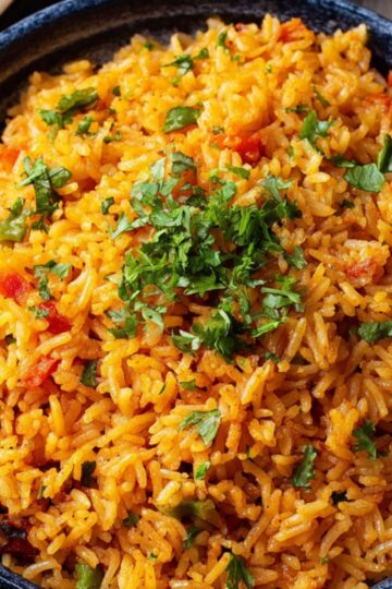 A vibrant tawa pulao of rice with cilantro and garnished with chopped tomatoes, served alongside lime wedges, suggesting a flavorful and fresh rice dish, possibly pilaf or seasoned rice.