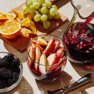 A bright kitchen setup with a glass being filled with red sangria, surrounded by fresh fruit like apples, oranges, grapes, and blackberries.