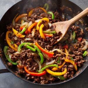 A pan of browned ground beef mixed with colorful bell peppers and onions being stirred with a wooden spoon.