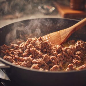 Ground beef being browned in a skillet with a wooden spatula, releasing steam as it cooks.