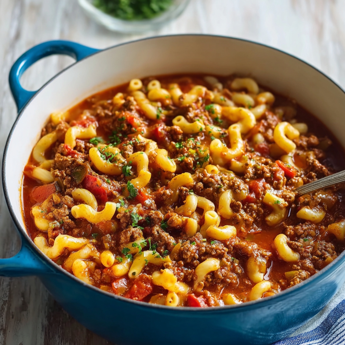 A pot of cooked American goulash in a blue Dutch oven, with tender macaroni, ground beef, and tomato sauce garnished with fresh parsley.