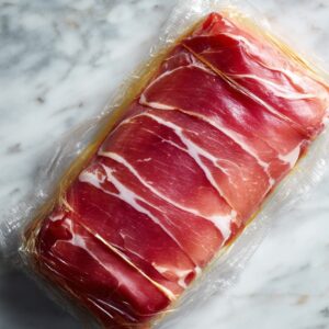 A person lays down plastic wrap on a counter and arranges slices of prosciutto for wrapping around the beef for Beef Wellington.