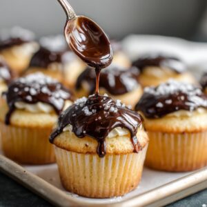 A spoon drizzling smooth chocolate ganache over freshly baked cream-filled cupcakes sprinkled with salt.