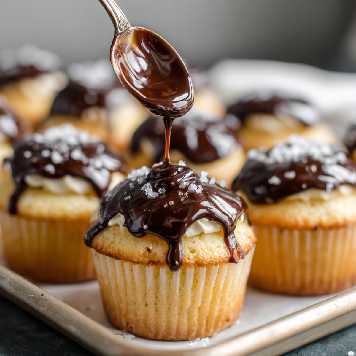 A spoon drizzling smooth chocolate ganache over freshly baked cream-filled cupcakes sprinkled with salt.