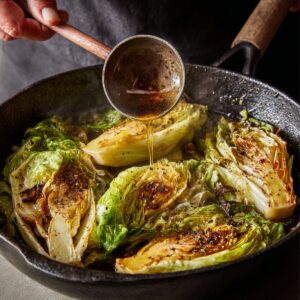 Cabbage wedges in a skillet being braised, while broth is ladled over the top for flavor.