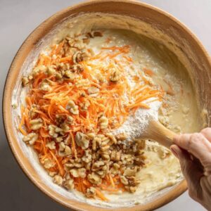 Mixing carrots and walnuts into batter: Wooden bowl filled with batter, grated carrots, and chopped walnuts being folded together.
