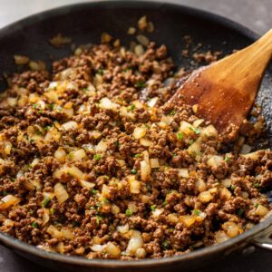 Browning the beef: Skillet with ground beef and diced onions cooking together, stirred with a wooden spatula.