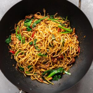 A wok with noodles being prepared by pouring in a prepared sauce, with steam rising.