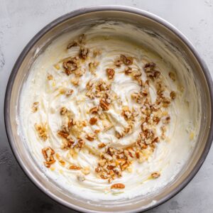 A bowl containing a whipped creamy frosting mixed with crushed pecans, ready to be used to decorate a cake with a soft, nutty touch.