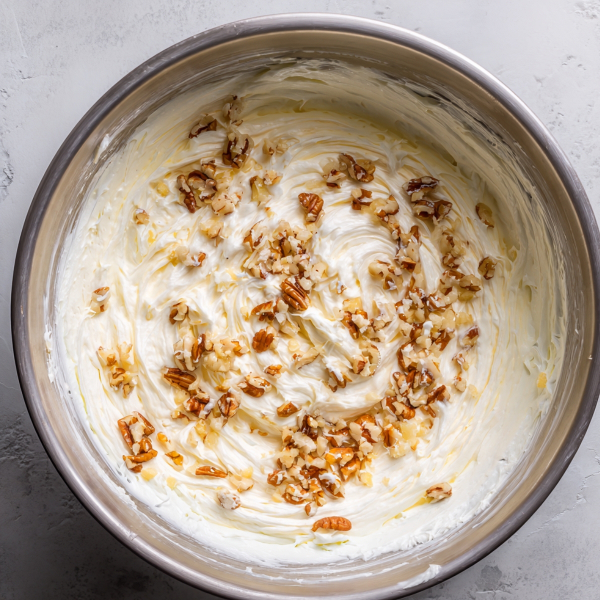 A bowl containing a whipped creamy frosting mixed with crushed pecans, ready to be used to decorate a cake with a soft, nutty touch.