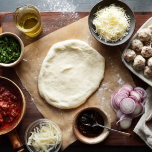 A pizza station is being prepared with various ingredients like olive oil for brushing, alongside the dough and other toppings, while the oven is preheated to 375°F.