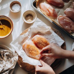 Overhead view of raw chicken breasts being seasoned with spices, salt, and pepper beside bowls of marinade and seasoning mix.