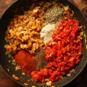 A pan showing diced onions, various spices, and tomatoes being cooked together for a curry base.