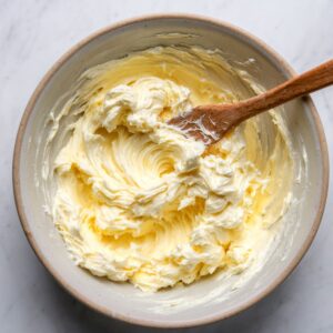 A bowl of creamy butter that has been mixed to a smooth consistency with a wooden spoon, placed on a marble countertop.