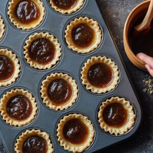 A muffin tin filled with unbaked tart shells, each with a dollop of brown sugar filling inside, ready to be baked.