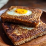 Two square slices of scrapple cooked with eggs in the center, stacked on a wooden plate with a golden-brown crust.