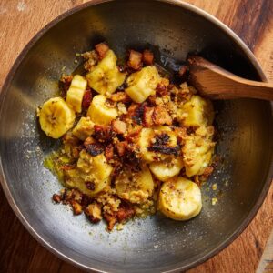 Sliced bananas, being sautéed with some bread or crouton pieces, creating a golden, crispy mixture.