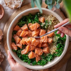 A bowl of quinoa and kale topped with cubed fresh salmon, seasoned lightly, with hands holding a spoon to mix the ingredients.