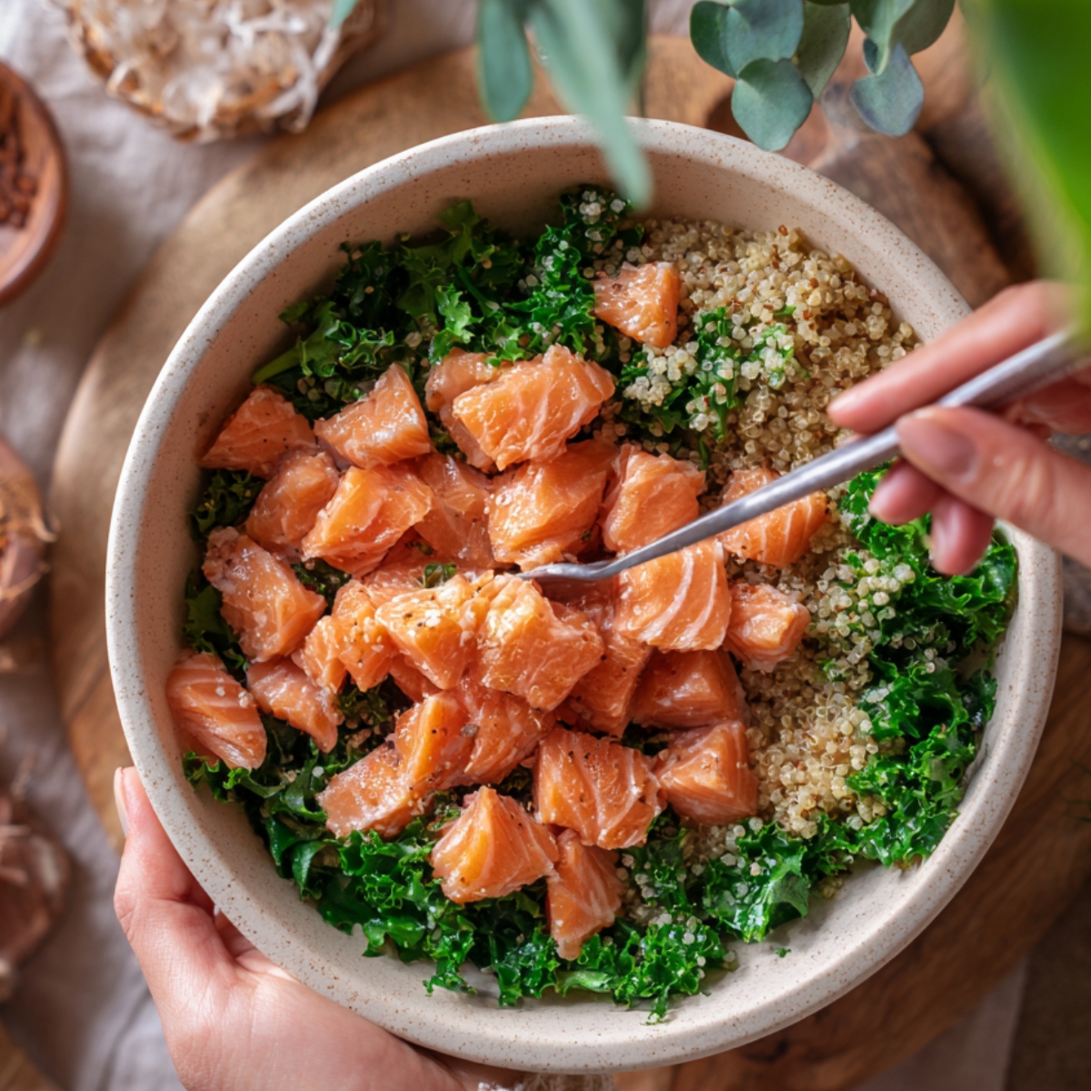 A bowl of quinoa and kale topped with cubed fresh salmon, seasoned lightly, with hands holding a spoon to mix the ingredients.