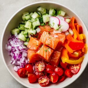 Colorful poke bowl featuring cubed raw salmon, cherry tomatoes, cucumber, red onion, bell peppers, and radishes arranged in a bowl.