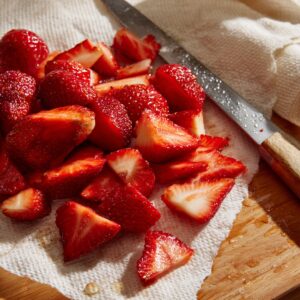 Freshly cut strawberries on a wooden surface, resting on a white cloth, with a knife nearby — glistening with moisture under natural light.