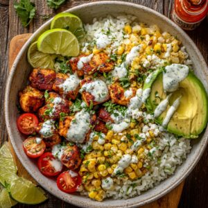 A Street Corn Chicken Rice Bowl with grilled chicken, roasted corn, avocado, lime wedge, and cilantro over white rice, drizzled with creamy sauce.