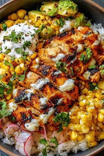 A Street Corn Chicken Rice Bowl with grilled chicken, roasted corn, avocado, lime wedge, and cilantro over white rice, drizzled with creamy sauce.