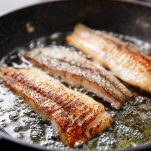 Several fish fillets are frying in a pan with sizzling oil, creating a crispy, golden brown texture on the fish.