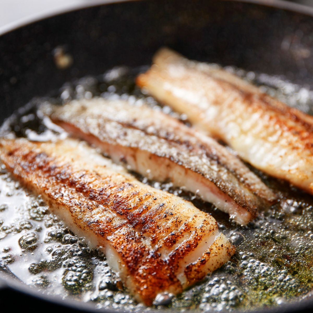 Several fish fillets are frying in a pan with sizzling oil, creating a crispy, golden brown texture on the fish.