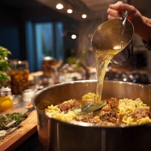 A kitchen scene with someone pouring broth into a pot of lasagna soup containing noodles, sausage, and herbs, surrounded by cooking ingredients.