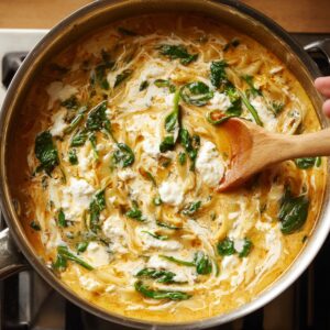 A pot of creamy lasagna soup simmering on the stove, showing wilted spinach, ricotta, and noodles swirled together with a wooden spoon.
