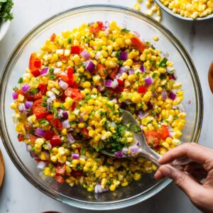A spoon mixing fresh corn salsa in a large glass bowl, filled with corn kernels, diced tomatoes, red onion, jalapeño, and chopped cilantro. Surrounding the bowl are ingredients like sliced red onion, cherry tomatoes, and chopped cilantro.