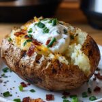 Air Fryer Baked Potato A loaded baked potato with a crispy skin, filled with fluffy potato and topped with sour cream, melted cheese, bacon bits, and chopped chives, served on a white plate with extra toppings scattered around.