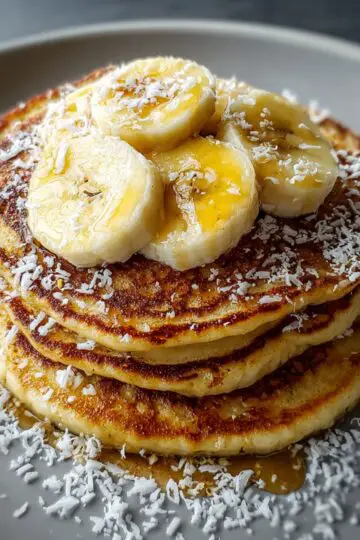 Banana Pancakes Another close, mouthwatering view of a pancake stack topped with fresh banana slices and syrup. The pancakes have crisp, caramelized edges and a soft, airy interior. Shredded coconut is sprinkled generously over the plate, and the warm lighting makes the syrup glisten. The gray plate and dark background create a beautiful contrast with the golden tones.