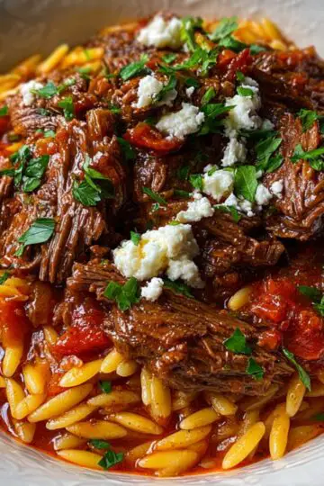 A hearty serving of Beef Giouvetsi, featuring succulent beef pieces simmered in a tomato and herb sauce with orzo pasta. The dish is garnished with fresh parsley and served in a white bowl over a wooden table. Each bite combines the deep, savory flavor of braised beef with the soft texture of perfectly cooked orzo — a comforting Mediterranean classic.