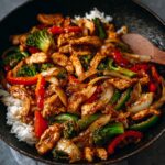 Chicken stir fry served over white rice in a black skillet, featuring broccoli, bell peppers, onions, and tender chicken coated in a rich savory sauce.