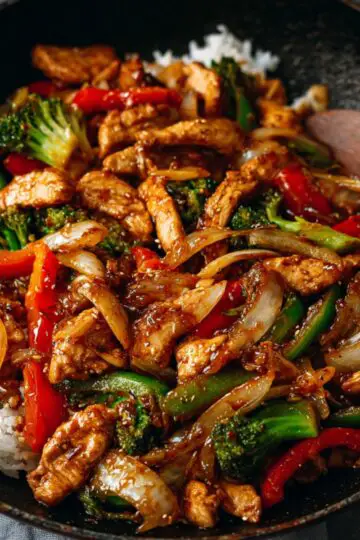 Chicken stir fry served over white rice in a black skillet, featuring broccoli, bell peppers, onions, and tender chicken coated in a rich savory sauce.