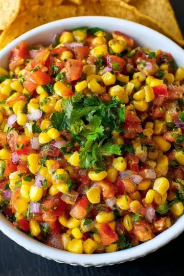 Corn Salsa Recipe A bowl of vibrant corn salsa filled with yellow corn, diced tomatoes, chopped red onion, cilantro, and jalapeño, garnished with fresh cilantro leaves, with tortilla chips in the background.