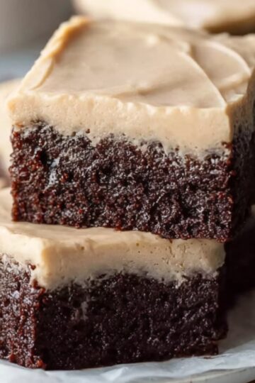 Frosted coffee brownies on a plate, showcasing dense chocolate layers topped with creamy coffee frosting, with a cup of coffee in the background.